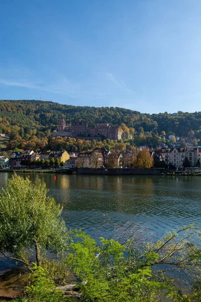 Arka planda bir tepenin üzerinde şatosu olan bir nehrin manzarası, sonbahar yaprakları ve nehir kıyısı boyunca renkli binalarla çevrili.