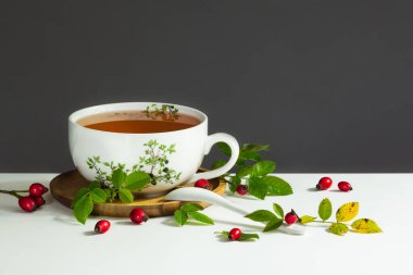 Beyaz çay kaşığı, kırmızı gül kalçası ve yeşil yapraklı bir fincan sağlıklı kalça çayı. Koyu gri arkaplan.
