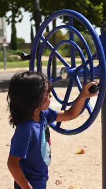 Uzun saçlı, spor yapan ve halka açık bir parkta spor aletleriyle oynayan bir çocuk.