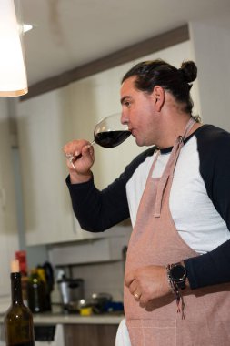 Ev mutfağında önlük giyerken bardaktan kırmızı şarap içen bir adam.