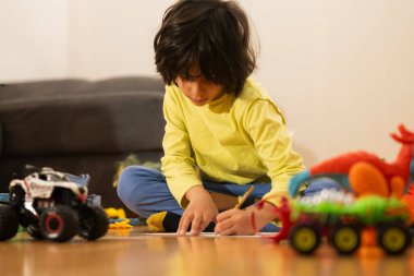 Küçük çocuk tahta zeminde oyuncaklarla otururken kağıda odaklanıyor.