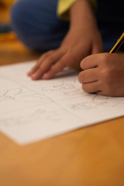Çocuğun elleri bir kalem tutuyor, çerçevelere bölünmüş beyaz bir kağıt üzerinde bir çizim yapıyor.