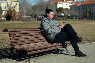 Parktaki bankta dinlenen, akıllı telefonuna bakıp sigara içen bir adam.