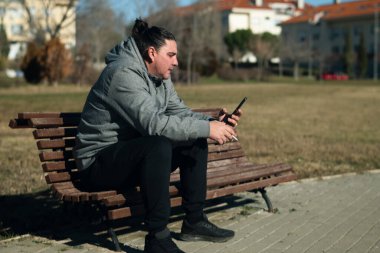 Parktaki bankta oturan bir adam elinde sigara tutarken akıllı telefonuna bakıyor.