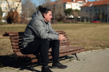 Parktaki bankta oturan genç bir adam, elinde bir sigarayla,