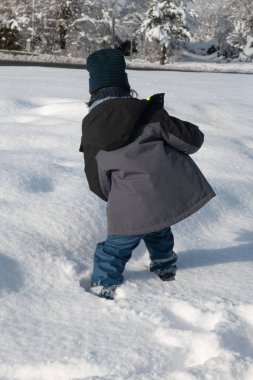 Genç bir çocuk güneşli bir günde kış kıyafetleri içinde taze kar üzerinde yürüyor.