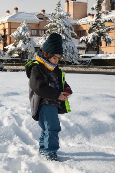 Karların içinde duran, elinde bir kartopu tutan, kışlık giysiler giyen genç bir çocuk.