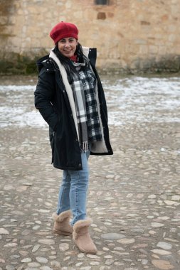 Kışlık palto giyen genç bir kadın, kırmızı bere, ekose atkı, kot pantolon ve botlar, dışarıda duruyor.