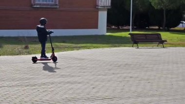 Güneşli bir günde şehir parkında kaldırımlı bir yolda modern elektrikli scooter süren bir çocuk.