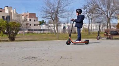 Güneşli bir günde, şehir parkında, kaldırımlı bir yolda, e-scooter kullanan bir çocuk.