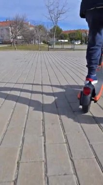 Asfaltlı bir şehir parkında elektrikli scooterını sürerken kask takan bir çocuğun arka görüntüsü.