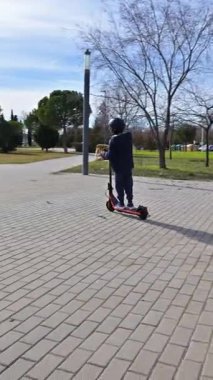 Güneşli bir günde şehir parkında elektrikli scooter kullanan kasklı bir çocuk.