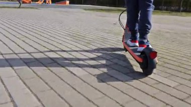 Güneş ışığı altındaki bir şehir parkında, kaldırımlı bir yol boyunca hareket eden bir e-scooter 'ın ayak görüntüsü.