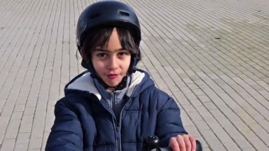 Uzun saçlı, koruyucu bir kask takan, kaldırımlı bir yolda e-scooter süren genç bir çocuk.