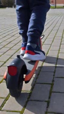 Kasklı genç bir çocuğun şehir parkındaki kaldırımlı bir yolda e-scooter sürüşünün arka görüntüsü.