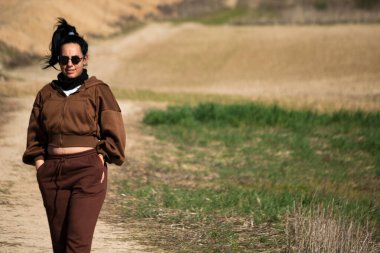 Güneş gözlüklü ve eşofmanlı bir kadın kırsal bir yolda dışarıda yürüyor.