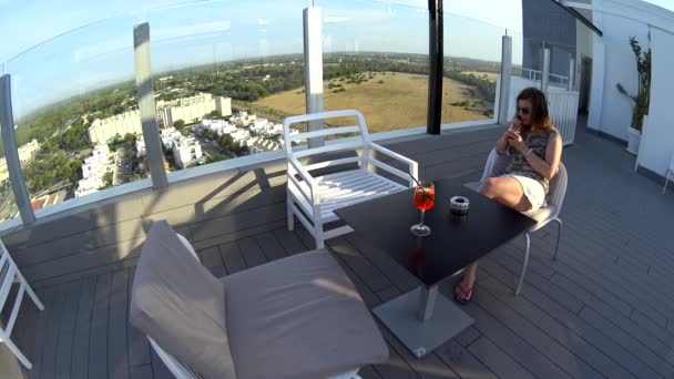 Fröhliches Mädchen in Sonnenbrille und Rock, das Cocktail auf Dachterrasse trinkt — Stockvideo