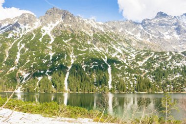Güneşli bir baharda Alp Gölü. Polonya 'nın yüksek tatra dağlarında, Rysy Tepesi' nin gezisi. Kayaların ve ladin ormanlarının arasındaki kar yamaçtaki suda yansıyor. Bulutlu mavi gökyüzü