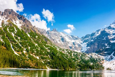 Tatra Ulusal Parkı 'ndaki Dağ Gölü. Morskie Oko ilkbaharın başlarında engebeli kar tepeleriyle çevriliydi. Su üzerindeki yansıma ve buzun güzel manzarası. yeşil arkaplan mavi gökyüzünün altında