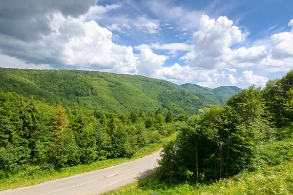Yazın dağ manzarası boyunca yol. Yukarıdan manzara. Bulutlu gökyüzünün altında, benekli ışıkta yeşil doğa manzarası. Ormanların arasında kıvrılan güzel vadi