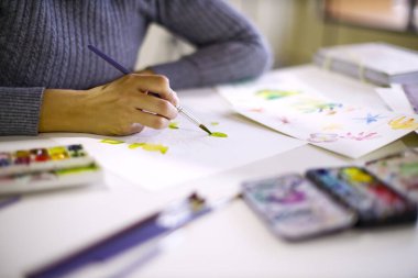 Beyaz kağıt üzerinde yeşil suluboya yaprakları boyayan bir fırça tutan sanatçı el, sanat malzemeleriyle birlikte beyaz bir masada yaratıcılık ve sanatsal ifade sergiliyor.
