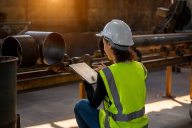 Koruyucu üniformalı genç bir kadın endüstriyel makineleri denetliyor ve fabrikadaki dijital tablet üzerinde gerekli notları alıyor..