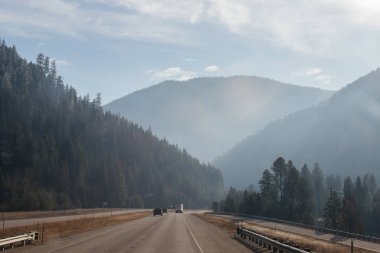 Arabaların ve kamyonların gittiği asfalt bir yol, dağların arasında, ufukta sisli dağlar, gökyüzü bulutlarla kaplı. Idaho, ABD, 11-23-2019