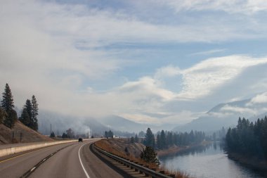 Güneşli bir günde güzel bir manzara, dağlar arasında bir otoyol, yüksek dağların önünde bulutlar ve sis, arabalar ve kamyonlar yol boyunca ilerliyor. Montana, ABD, 11-23-2019