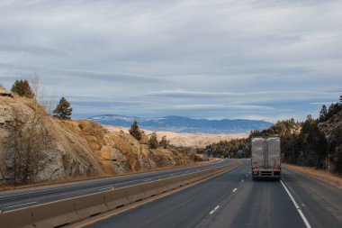 Bir sonbahar gününde yol manzarası, kenarlarında ağaçların, kamyonların sürdüğü ağaçların, uzaklardaki dağların ve ağaçların büyüdüğü dik kayaların olduğu bir otoyol..