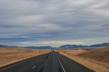 Bozkırların arasında asfalt yol, sarı kuru çimenler ufukta mavi dağlar, güzel mavi-gri sonbahar gökyüzü. Montana, ABD, 11-23-2019