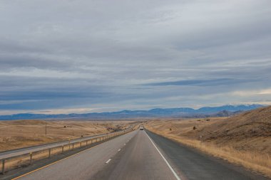 Bozkırların arasında asfalt yol, sarı kuru çimenler ufukta mavi dağlar, güzel mavi-gri sonbahar gökyüzü. Montana, ABD, 11-23-2019