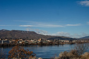 Geniş bir nehrin ardında güneşli bir günde mavi dağların arka planına karşı parlak mavi gökyüzünün arka planına karşı bir şehir.