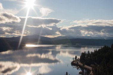 Büyük gölü olan güzel bir manzara, gölü çevreleyen mavi dağlar, açık mavi gökyüzü ve güneş.