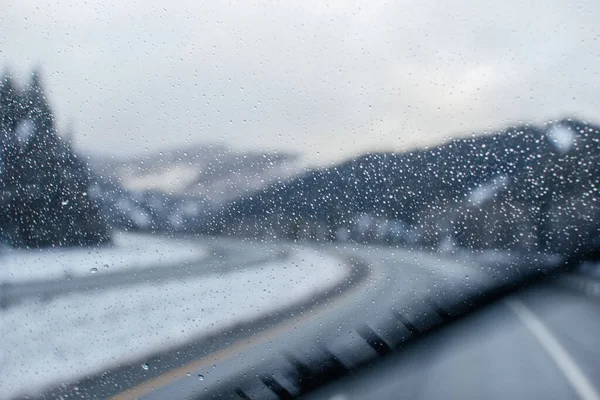 Car glass fogged up with rain drops, through which a blurred highway among snowy mountains is visible
