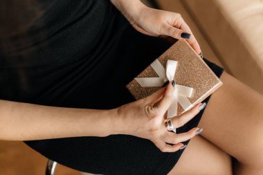 Güzel siyah ve gri manikürlü kısa siyah elbiseli bir kız bej renkli şirin kurdeleli parlak altın bir hediye kutusu tutuyor.