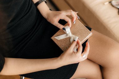 Güzel siyah ve gri manikürlü kısa siyah elbiseli bir kız bej renkli şirin kurdeleli parlak altın bir hediye kutusu tutuyor.