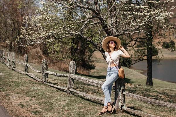 A beautiful girl with brown curly hair, sunglasses and a straw hat, linen cropped blouse with a knot and jeans, stands near an old fence under flowering dogwood trees on a sunny spring day.