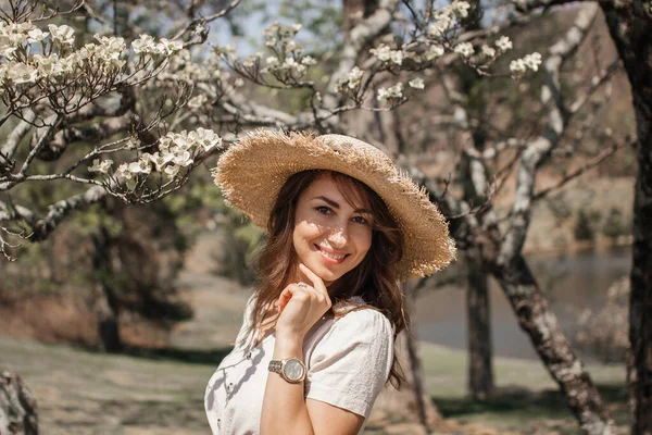 A beautiful girl with brown curly hair, sunglasses and a straw hat, linen cropped blouse with a knot and jeans, stands near an old fence under flowering dogwood trees on a sunny spring day.