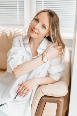 Beyaz tişörtlü ve kot pantolonlu güzel sarışın bir kız bej renkli bir kanepede güneşli bir odada oturuyor.