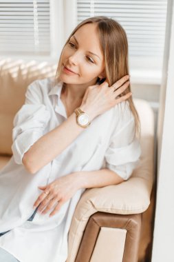 Beyaz tişörtlü ve kot pantolonlu güzel sarışın bir kız bej renkli bir kanepede güneşli bir odada oturuyor.