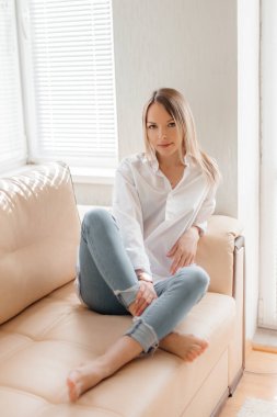 Beyaz tişörtlü ve kot pantolonlu güzel sarışın bir kız bej renkli bir kanepede güneşli bir odada oturuyor.