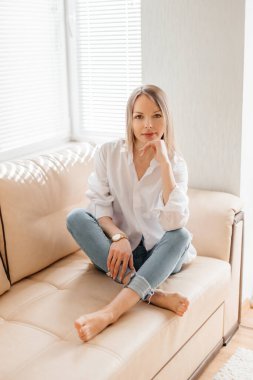 Beyaz tişörtlü ve kot pantolonlu güzel sarışın bir kız bej renkli bir kanepede güneşli bir odada oturuyor.