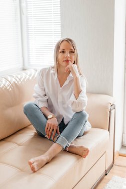 Beyaz tişörtlü ve kot pantolonlu güzel sarışın bir kız bej renkli bir kanepede güneşli bir odada oturuyor.