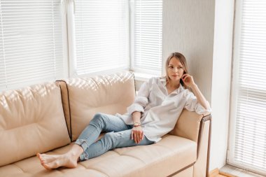 Beyaz tişörtlü ve kot pantolonlu güzel sarışın bir kız bej renkli bir kanepede güneşli bir odada oturuyor.