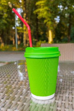 Parktaki cam bahçe masasında kapaklı ve kırmızı pipetli parlak yeşil kağıt bardak. Arka planda bulanık yeşil ağaçlar açık hava havası yaratır.