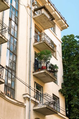 Pastel renkli bir binanın ön cephesi alçak açılı balkonları ve dikey metal drenaj boruları var. Bir balkondaki yeşil saksı bitkileri mimari bileşimine doğal bir aksan ekler.
