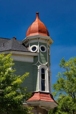 Bu çarpıcı mimari detay, yıpranmış kırmızı-turuncu konik çatılı ve belirgin yuvarlak pencereli belirgin bir kuleye sahip tarihi bir konutun üst kısmını yakalar. Açık yeşil pano kaplaması karışık bir beyazla süslenmiş.