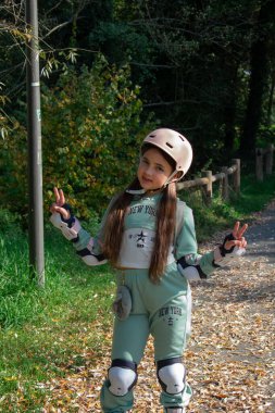 8 yaşındaki neşeli bir kız, renkli yapraklarla dolu bir sonbahar parkında paten kayıyor. Kask ve koruyucu giysiler giyerek aktif ve neşeli bir açık hava gününün tadını çıkarıyor..