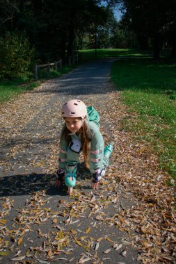 8 yaşındaki neşeli bir kız, renkli yapraklarla dolu bir sonbahar parkında paten kayıyor. Kask ve koruyucu giysiler giyerek aktif ve neşeli bir açık hava gününün tadını çıkarıyor..