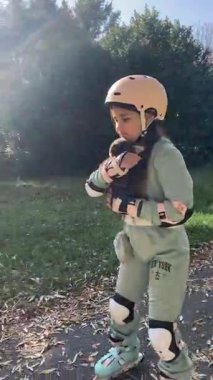 8 yaşındaki neşeli bir kız, renkli yapraklarla dolu bir sonbahar parkında paten kayıyor. Kask ve koruyucu giysiler giyerek aktif ve neşeli bir açık hava gününün tadını çıkarıyor..